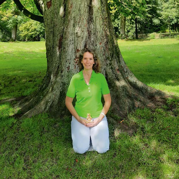 Myriam Ingenfeld kniet ruhig am Fuß eines großen Baumes in einem Park